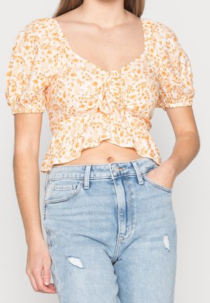 Vrouw die een gele korte top met bloemen en gepofte mouwen draagt, gecombineerd met lichtblauwe gescheurde jeans, hand in de zak, torso zichtbaar.