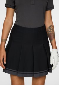 Persoon in een zwarte geplooide rok met donkergrijze strepen, een donker polohemd, een witte golfhandschoen en zichtbare tatoeages op de armen.