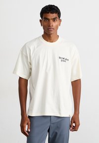 Cream-colored cotton t-shirt with short sleeves and a loose fit. Features a black logo on the chest area. Smooth texture.