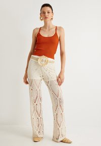 Top arancione a righe con spalline sottili abbinato a pantaloni di pizzo bianchi con motivi a diamante. Il modello indossa una cintura intrecciata con una fibbia floreale.