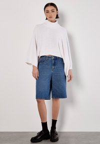 Weißer, gestrickter, kurzer Rollkragenpullover mit weiten Ärmeln; kombiniert mit blauen Jeans-Bermudashorts und schwarzen Plateauschuhen.