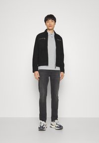 Sort ruskindsjakke med lynlåslommer, grå polo-shirt, mørke jeans og multikolor sneakers, præsenteret mod en ensfarvet baggrund.