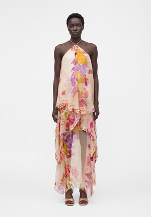 Kvinde iført en ærmeløs, lagdelt, blomsterprintet halterneck-maxikjole med lyserøde, lilla, gule og røde mønstre og beige åbne hæle.