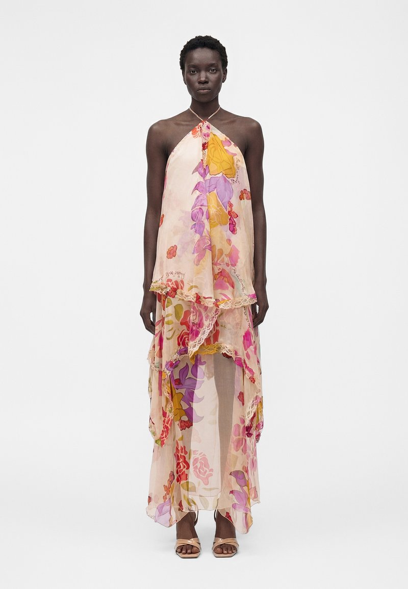 Γυναίκα που φορέσει ένα αμάνικο, στρωματοποιημένο, φλοράλ μάξι φόρεμα με ροζ, μωβ, κίτρινα και κόκκινα σχέδια και μπεζ ανοιχτά παπούτσια με τα δάχτυλα έξω.