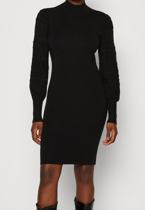 Schwarzes, knielanges, figurbetontes Kleid mit hohem Kragen und strukturierten, langen Puffärmeln, kombiniert mit schwarzen Stiefeln.