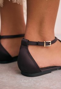 Chaussures plates marron avec une finition lisse semblable à du cuir. Dotées d'une fine sangle pour la cheville avec une boucle dorée. Design élégant et profil bas.