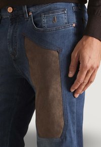 Jeans in denim con una finitura blu liscia, caratterizzati da un'etichetta in suede marrone sulla coscia e un classico design a cinque tasche.