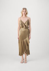 Olivgrön satinklänning med spaghettiband, volangkantad bodice och asymmetrisk fåll. Har en flödande silhuett och en subtil lyster.