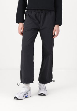 Person trägt schwarze zulaufende Hosen mit Kordelzügen an den Knöcheln, weiße Socken und weiße Laufschuhe mit blauen Akzenten.