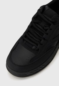 Black athletic shoe with a smooth synthetic leather upper, featuring a rounded toe, textured accents, and black laces. Rubber sole included.