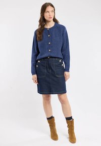 Cardigan en tricot bleu marine avec des boutons en bois, associé à une jupe en denim foncé avec des accents dorés et des poches, complété par des bottines beige.