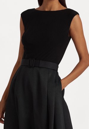 Kobieta w czarnej sukience bez rękawów z dopasowanym żywotem, szerokim paskiem z prostokątną klamrą i rozkloszowaną spódnicą, z ręką w kieszeni.