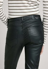 Pantalones de imitación de cuero negros con un diseño ajustado, que cuentan con bolsillos traseros y detalles de costura visibles. Parte de una blusa a rayas es parcialmente visible.