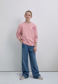 Pink sweatshirt with a graphic logo on the chest, paired with wide-leg blue jeans featuring star patterns. The model wears patterned sneakers.
