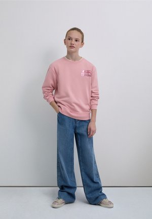 Sweat-shirt rose avec un logo graphique sur la poitrine, associé à un jean bleu à jambes larges orné de motifs d'étoiles. Le mannequin porte des baskets à motifs.