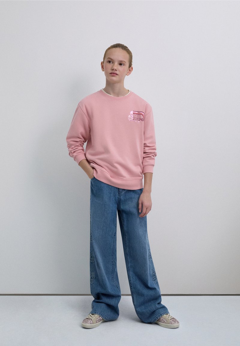 Pink sweatshirt with a graphic logo on the chest, paired with wide-leg blue jeans featuring star patterns. The model wears patterned sneakers.