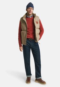 Braune gepolsterte Weste, roter Zopfmuster-Pullover, blaues Hemd, marinefarbene Mütze, dunkle Jeans, braune Schnürstiefel mit beigen Sohlen.