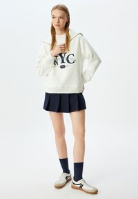 Sudadera blanca con capucha y gráfico de NYC en azul marino, combinada con una falda plisada azul marino, calcetines azul marino y zapatillas blancas con detalles en negro.