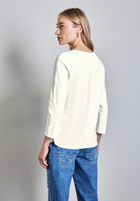 Top bianco a maniche lunghe in morbido tessuto con scollatura rotonda, indossato con jeans blu rilassati. Design semplice, con dettagli di cucitura delicati sulle maniche.
