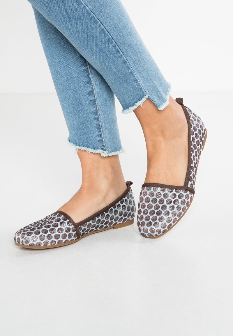 Des pieds portant un jean court bleu clair et des chaussures plates à enfiler avec des pois marron et une bordure sur fond blanc.
