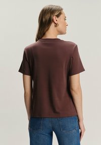 Kobieta ubrana w prostą, brązową koszulę z krótkim rękawem i niebieskie dżinsy, zwrócona tyłem, na jasnym, neutralnym tle.