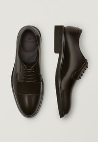 Tummanruskeat nahkakengät, joissa on sileä pinta. Niissä on nauhakiinnitys, pyöristetty varvas ja matala korko. Sivussa on sauman yksityiskohtia.