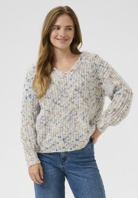 Vrouw met lang haar, gekleed in een gemêleerd gebreid vest en blauwe spijkerbroek, glimlachend, met één hand op haar heup tegen een effen achtergrond.
