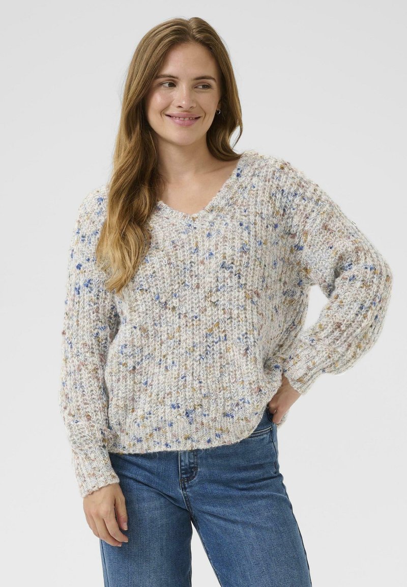 Vrouw met lang haar, gekleed in een gemêleerd gebreid vest en blauwe spijkerbroek, glimlachend, met één hand op haar heup tegen een effen achtergrond.