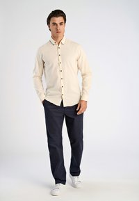 Lichtbeige button-up shirt met donkere knopen, gecombineerd met marineblauwe chinos en witte sneakers. Het shirt heeft een afgeronde onderkant en lange mouwen.