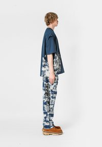 Joven de pie de lado, vistiendo un atuendo suelto de estampado abstracto en azul marino y blanco, con zapatos de plataforma marrones, sobre un fondo blanco.