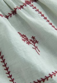 Tissu blanc à tissage texturé présentant une broderie florale rouge foncé et des coutures à motif de feuilles le long des coutures.