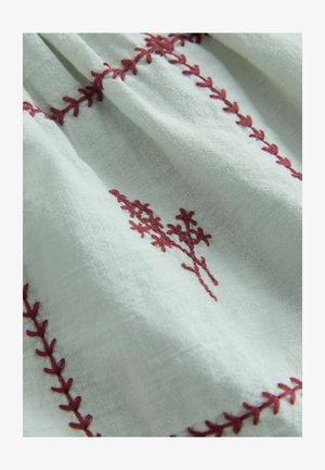 Bijela tkanina s teksturiranim tkanjem sadrži tamnocrveni cvjetni vez i šavove s uzorkom listova duž šavova.