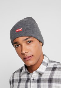 Grå strikket hue med et rødt Levi's logo-patch, der har en tætsiddende pasform og rillet tekstur. Bæres af en person med en rutete skjorte.