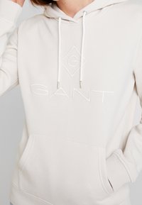 Vaaleabeige huppari, jossa on etutasku, kiristysnauha ja kohokuvioitu "GANT"-logo valkoisena. Sileä pinta, rento muotoilu ja resorit hihansuissa.