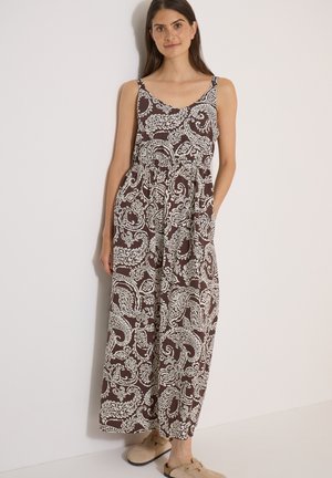 Kvinde står op ad en hvid væg iført en ærmeløs brun maxikjole med hvidt paisleymønster og beige slip-on sko.
