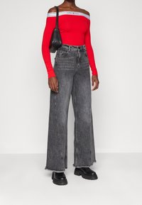 Nainen, jolla on päällään punainen olkaimeton paita, korkeavyötäröiset harmaat leveälahkeiset farkut, joissa on raakat hihat, mustat paksupohjaiset saappaat ja pieni musta olkachelmi.