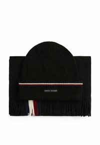 Conjunto de gorro de punto negro y bufanda a juego. La bufanda tiene flecos con borlas. El gorro presenta una raya contrastante y una etiqueta de logo. Tejido texturizado.
