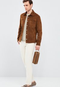 Bruine suede jas met een overhemdkraag, twee borstzakken, zilveren drukknoopjes, gecombineerd met crème broek en een bruine leren tas.
