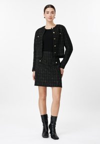 Γυναίκα που φοράει μαύρο καρό σακάκι με κουμπιά και ταιριάζουσα φούστα, μαύρο τοπ και μαύρα μποτάκια μέχρι τον αστράγαλο, στέκεται μπροστά σε λευκό φόντο.