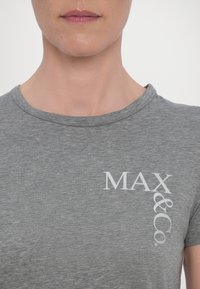Gros plan d'une personne portant un T-shirt gris avec un texte blanc "MAX&Co." sur le haut de la poitrine, montrant le cou et le bas du visage.