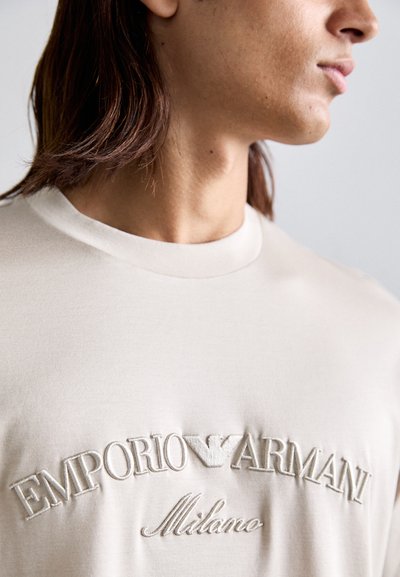 Camiseta de algodón beige con el logotipo bordado "EMPORIO ARMANI Milano" en el pecho. Diseño de cuello redondo con una textura suave.