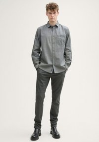 Chemise à boutons grise avec une poche sur la poitrine, associée à un pantalon tapered gris foncé et des bottines noires, présentée sur un fond neutre.