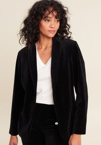 Blazer in corduroy nero con colletto a scialle e maniche lunghe, abbinato a una maglietta bianca a scollo a V. Presenta righe verticali e una vestibilità morbida.