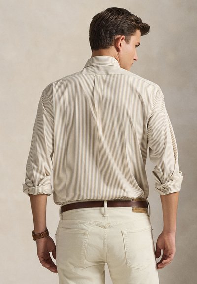 Μπεζ και λευκό ριγέ πουκάμισο με κουμπιά και μανίκια γυρισμένα, παρουσιασμένο σε ανδρικό μοντέλο, συνδυασμένο με ανοιχτόχρωμο τζιν και καφέ ζώνη.