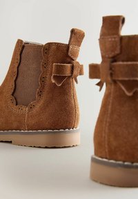 Botines de ante marrón con bordes festoneados y acentos de lazo decorativos, que cuentan con inserciones elásticas en los laterales y una suela de goma texturizada.
