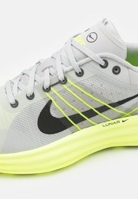 Chaussure de sport gris clair avec des accents noirs et jaunes. Comprend une tige en mesh, des motifs texturés et une semelle épaisse jaune néon.