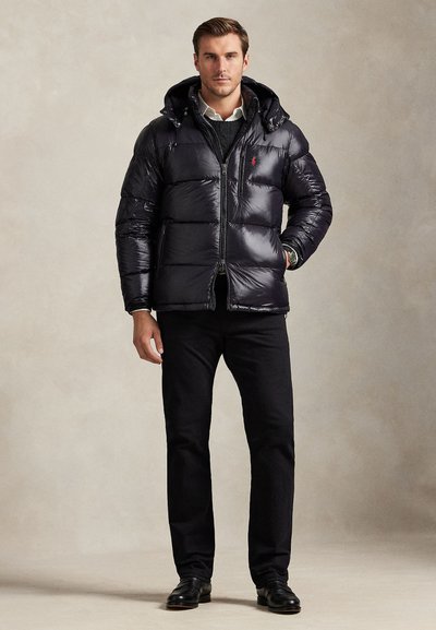 Man wearing a black shiny puffer jacket with a hood, black pants, and black shoes standing against a plain beige background.