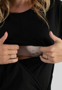 Schwarzes Oberteil mit einer drapierten Öffnung, die einen hellbeigen, gemusterten BH darunter zeigt. Die Hände greifen das Material und heben den Schichteffekt hervor.