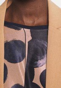 Beżowa marynarka o gładkim, teksturowanym wykończeniu, zestawiona z topem bez rękawów z niebiesko-różowym okrągłym wzorem na jasnym tle.