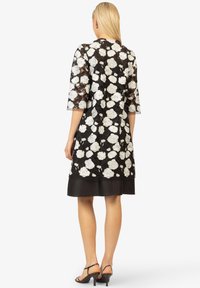 Schwarzes Spitzenkleid mit weißen Rosenmustern, dreiviertel Ärmel, glatte schwarze Fütterung und ein ausgestellter Saum über dem Knie.